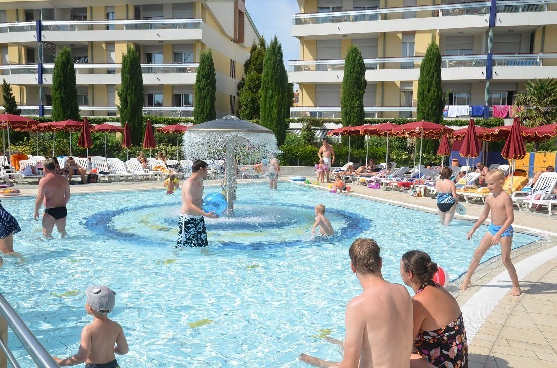 Rodiny s dětmi u bazénu s vodní atrakcí ve Villaggio Planetarium Resort v Bibione v Itálii, ideální místo pro rodinnou dovolenou u moře.